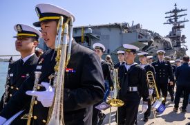 About a dozen South Korean service members walk in a line in formal dress uniforms and holding instruments, mostly walking two by two. Behind them, an aircraft carrier looms in the harbor against a blue sky.