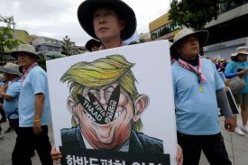A close-up view shows several protesters in a crowd beneath a pale gray cloudy sky. Many of the demonstrators wear hats and light blue shirt. The person closest to the camera holds up a sign showing a political cartoon-style image of Donald Trump with missiles sticking out of his mouth, with writing in Korean.
