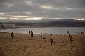 Five men play soccer on a beach in Spain. beneath a cloudy sky, low mountains visible nearly touching the clouds in the distance. The men are wearing short and T-shirt and are barefoot as they kick the ball around the sand.