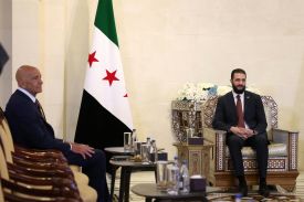 Sharaa sits in an ornately decorated armchair with his hands folded in his lap. He is in his young 40s, wearing a suit and a beard. Barrack, an older bald man who also wears a suit, sits in another armchair at a diagonal. They face small low tables with water glasses on them; a Syrian flag hangs in the corner.