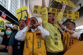 About half a dozen people are seen at the front of a crowd, most of them wearing yellow and holding yellow signs with writing in Mandarin. One person holds their hands up to their face like a megaphone as they shout; others wave their hands in the air.