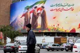 A man in a black jacket and beanie walks underneath a banner with an illustration displaying a transfer of the Iranian flag between the three supreme Leaders.