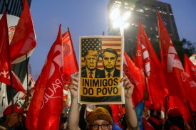 A person with glasses holds up a sign with Trump and Bolsonaro's faces, and the text: "INIMIGOS DO POVO." The person is surrounded by people holding up red flags.