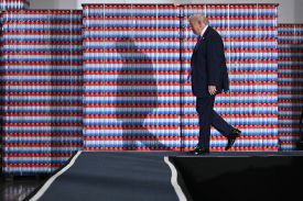 Trump walks across a stage in front of a wall made up of stacks of red, white, and blue drink cans, which rise up to a height just above his head.
