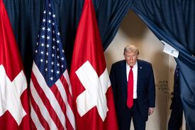 Trump dressed in a suit and red tie walks through a blue-curtained doorway. In the foreground, U.S. and Swiss flags are shown standing side by side.