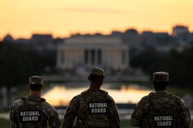 Three members of the National Guard are seen from behind as they stand in front of Washington's Reflecting Pool at dusk, the sky and the water lit up orange with the sunset. The Lincoln Memorial stands at the shore across the water. All three guard members wear green camouflage uniforms, matching hats, and bulletproof vests that say "National Guard" on their backs.