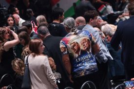A group of dozens of people of varying ages are seen mostly from behind. Close to the camera is a tall person wearing a red-white-and-blue shirt with photos of Orban's and Trump's faces printed on the back, along with text in English that says "Friendship matters—leaders of the free world."