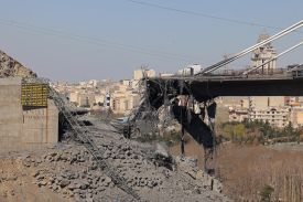 Beneath a hazy blue sky, a bridge stretches from the right of the image toward the left. The bridge crumbles into nothing before it reaches the left abutment, debris and rubble scattered underneath it. Midrise buidlings are visible beyond.