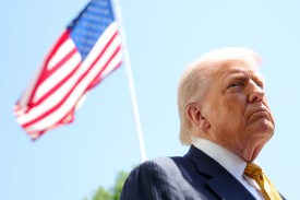 President Trump frowning slightly against a clear blue sky with an American flag out of focus behind him.
