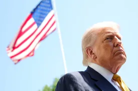 President Trump frowning slightly against a clear blue sky with an American flag out of focus behind him.