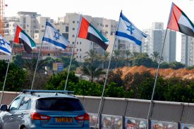 A car is seen from behind as it passes by beneath Israeli and United Arab Emirates flags flying on poles. In the background are midrise buildings and palm trees and other greenery.