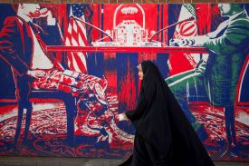 An Iranian woman wearing a black dress and black headscarf is seen in profile as she walks on a sidewalk in front of a brightly colored wall mural. The red-white-and-blue mural depicts two men seated at a table, identified with placards as representing the U.S. and Iran. The U.S. official wears military fatigues from the waist down, but a suit on his top half.