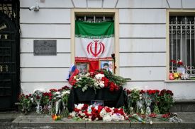 Red and white flowers spill out from a makeshift altar with a framed portrait of Ayatollah Ali Khamenei resting on a stoop outside a building. An Iranian flag hangs over the door above the stoop, and a smaller Russian flag is stuck among the flowers.