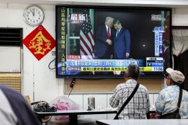 Two people are seen from behind stand with their necks craned to look up at a TV screen showing a news broadcast. On the screen, a video shows Trump and Xi shaking hands.