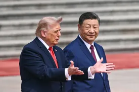 Two men in dark suits standing outdoors. Donald Trump turns slightly toward Xi Jinping with his mouth open and his palms held upward in a gesturing motion. He wears a bright red striped tie. Xi has a slight smile, looking forward. He wears a dark burgundy tie. The background consists of blurred gray stone steps and a red carpet.