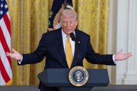 U.S. President Donald Trump stands in front of a podium bearing the U.S. presidential seal. He speaks into a microphone while holding up both hands.
