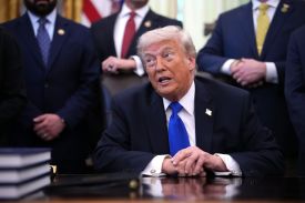 U.S. President Donald Trump sits behind his desk in the Oval Office with his hands clasped in front of him. He is speaking and looking off to the side. Three blurred figures wearing suits stand in the background.