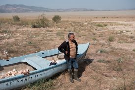 Rafet Kurse, a former fisherman, stands next to an abandoned boat on the former shores of Marmara Lake. A dry, dusty landscape stretches into the distance behind him.