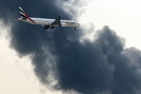An Emirates commercial jet flies through a plume of black smoke that covers up much of the sky, with just a few windows of light peeking through.