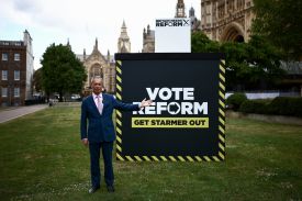 Reform UK leader Nigel Farage poses in front of a sign reading, “Vote Reform, Get Starmer Out.” He is wearing a suit, and his left arm is outstretched.