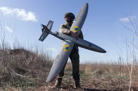 A Ukrainian soldier in camouflage holds a large, gray drone with a wingspan a little longer than he is tall, with yellow and blue markings on its wings. He's standing in a field of long, dry glass beneath a pale blue sky.