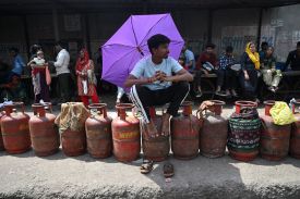A man wearing a T-shirt and track pants sits perched on a gas container, his sandals off and resting on the ground in front of him. He holds a purple umbrella over his head, seemingly to provide shade. Other people mill about in the background.