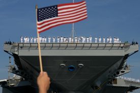 U.S. Navy sailors are seen from afar, standing in a line along the edge of the deck of the USS Gerald R. Ford aircraft carrier. From below, in the foreground, a hand waves a small American flag.
