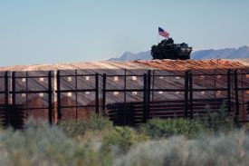 A military vehicle flies a U.S. flag from a hill overlooking a tall border fence with brush in the foreground.