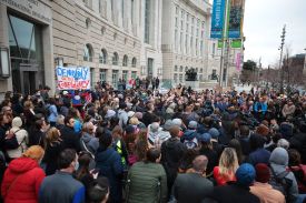 A large crowd of people, some holding aloft a banner that reads "Democracy died in complacency," gather outside the headquarters of the U.S. Agency for International Development on an overcast day in Washington to listen to speeches condemning the closure of the agency.
