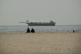 Three people sit on a beach while a vessel sails in the water.