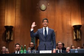 Warsh, standing in a dark business suit and tie, raises his right hand to swear an oath in a large chamber lit by wall sconces. Other people in formal attire sit behind him with bottles and glasses of water scattered on their desks.
