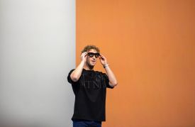 Mark Zuckerberg stands on a stage, adjusting a pair of thick-rimmed black smart glasses. He is wearing a black T-shirt and blue jeans, positioned against a background that is half white and half solid orange.