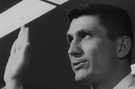 A man in a white collared shirt raises his right hand as if taking an oath or being sworn in. The image is a black-and-white, close-up shot of his profile.