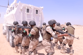 Soldiers hold guns as they stand behind an armored vehicle.
