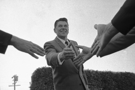 A black and white photograph from a low angle shows a smiling man in a suit reaching out to shake hands with several people. His arm is extended toward the camera, and the hands of three other people in suits enter the frame from the sides, reaching back toward him. The background features a clear sky and the top of a manicured hedge.